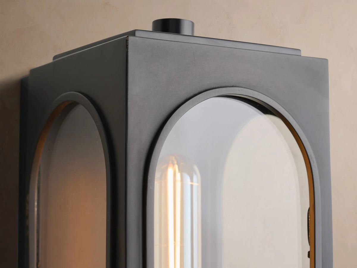 Palladian Outdoor Sconce. Black, rectangular light with arched glass windows & exposed filament bulb, mounted on a beige wall.