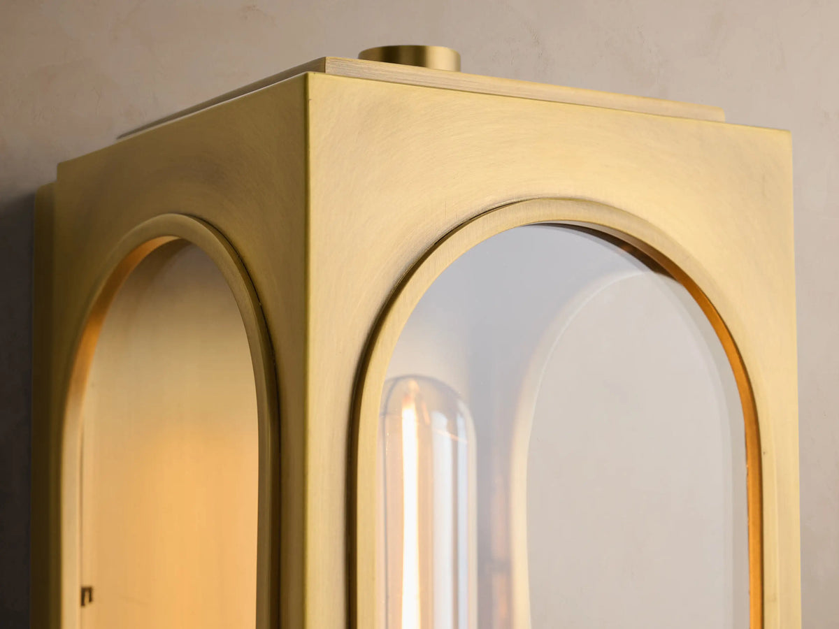 Palladian Outdoor Sconce: Vertical golden brass light with arched glass, unlit Edison bulb, mounted on a beige wall.