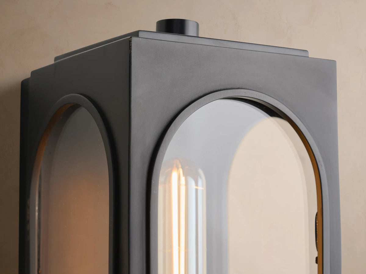 Dark gray Palladian Outdoor Sconce, rectangular with arched top, showcasing a golden tubular bulb against a beige wall.