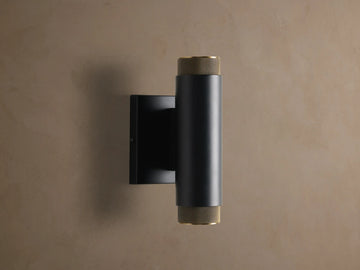 Matte black cylindrical Spotlight Outdoor Sconce with brass knurled end caps, mounted vertically on a light brown wall.