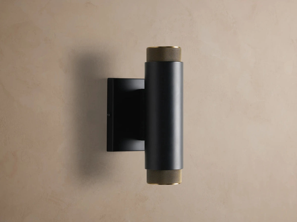 Matte black cylindrical Spotlight Outdoor Sconce with brass knurled end caps, mounted vertically on a light brown wall.