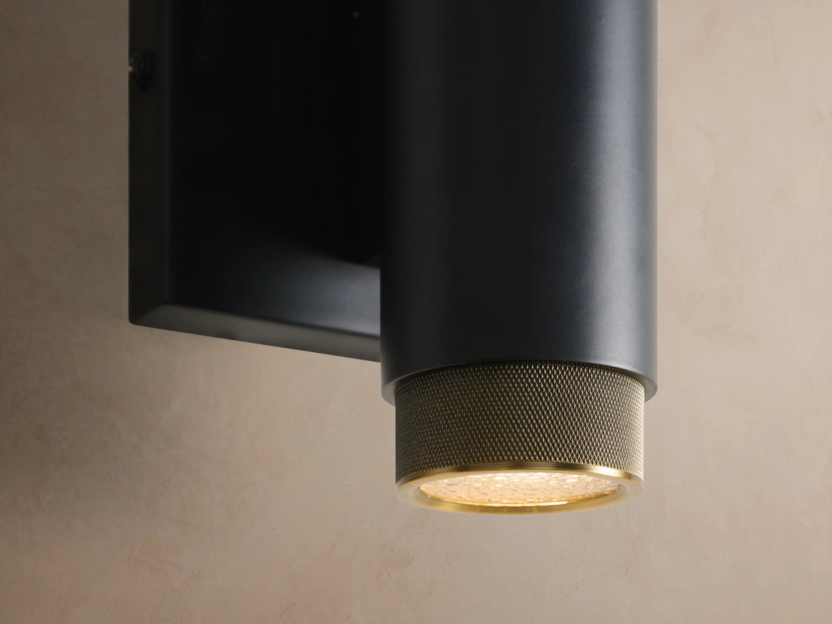 Matte black cylindrical Spotlight Outdoor Sconce with brass knurled ends, mounted horizontally on a light beige wall.