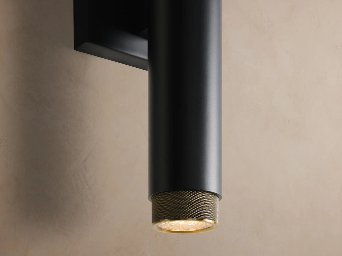 Matte black cylindrical Spotlight Outdoor Sconce with brass knurled end caps, mounted vertically on a light brown wall.