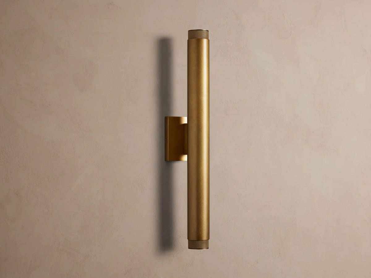 Vertical brass spotlight outdoor sconce with cylindrical body and knurled end caps, mounted on a neutral wall.