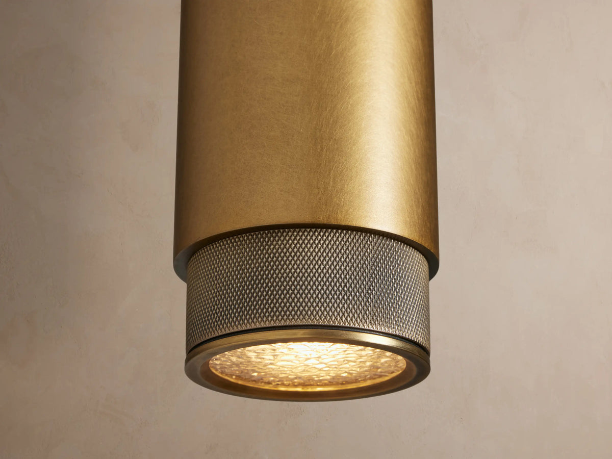 Vertical brass Spotlight Outdoor Sconce with a cylindrical body and knurled ends, mounted on a light beige wall, casting a shadow.