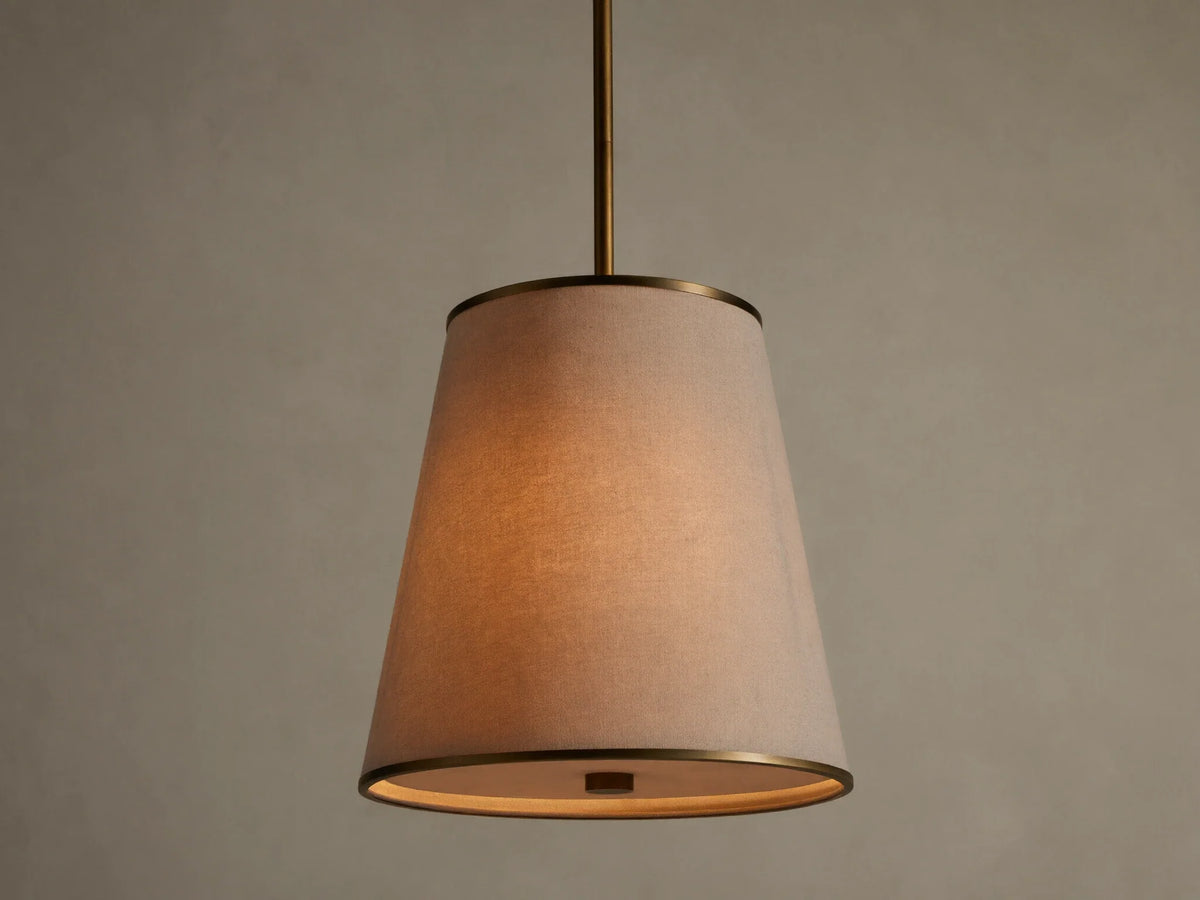 Adria Pendant with a light fabric conical shade, brass trim, hanging and emitting a warm glow against a textured wall.