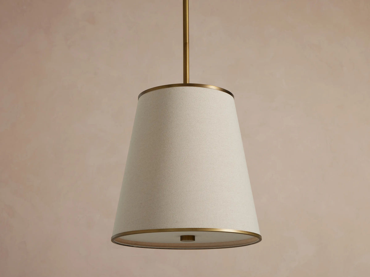 Adria Pendant with a light fabric conical shade, brass trim, hanging and emitting a warm glow against a textured wall.
