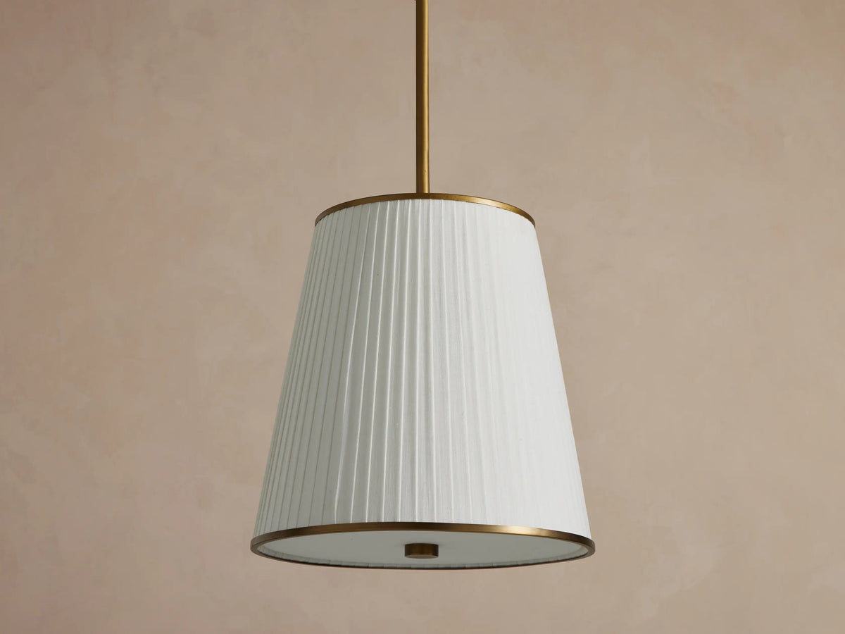 Adria Pendant: White pleated conical shade with brass trim, illuminated, hanging from a rod against a neutral wall.