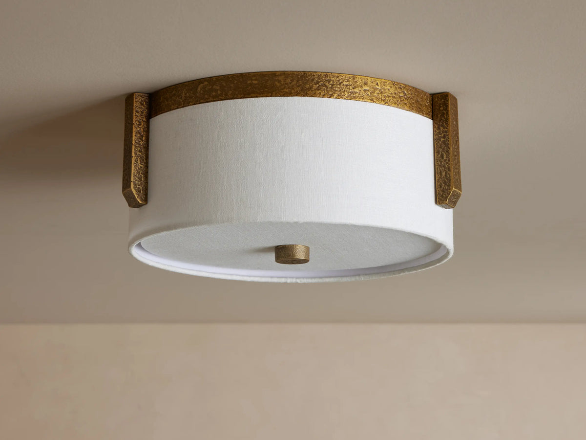 Emmet Flush Mount. Illuminated round white linen shade framed by textured antiqued brass, mounted on a light-colored ceiling.