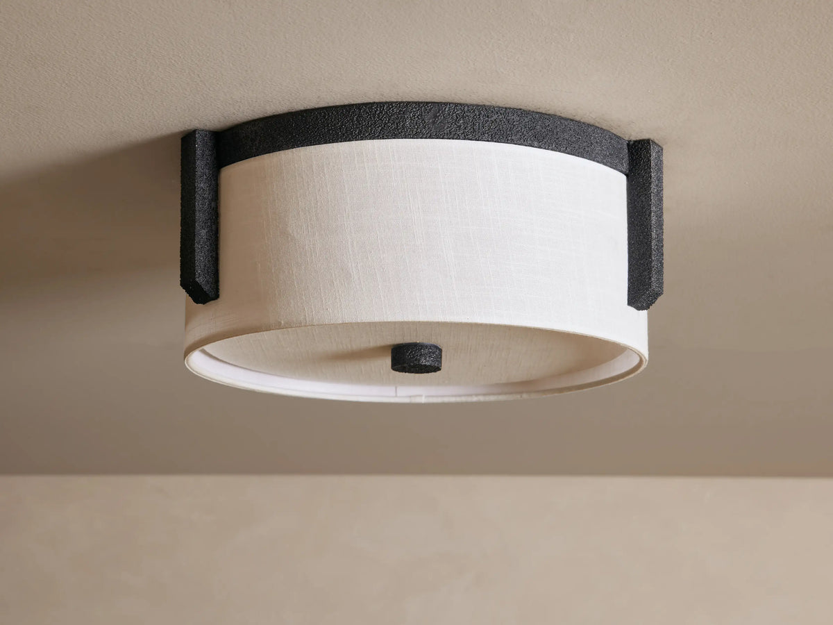 Emmet Flush Mount ceiling light with a glowing white linen drum shade and dark, textured sand-casted bronze frame on a ceiling.