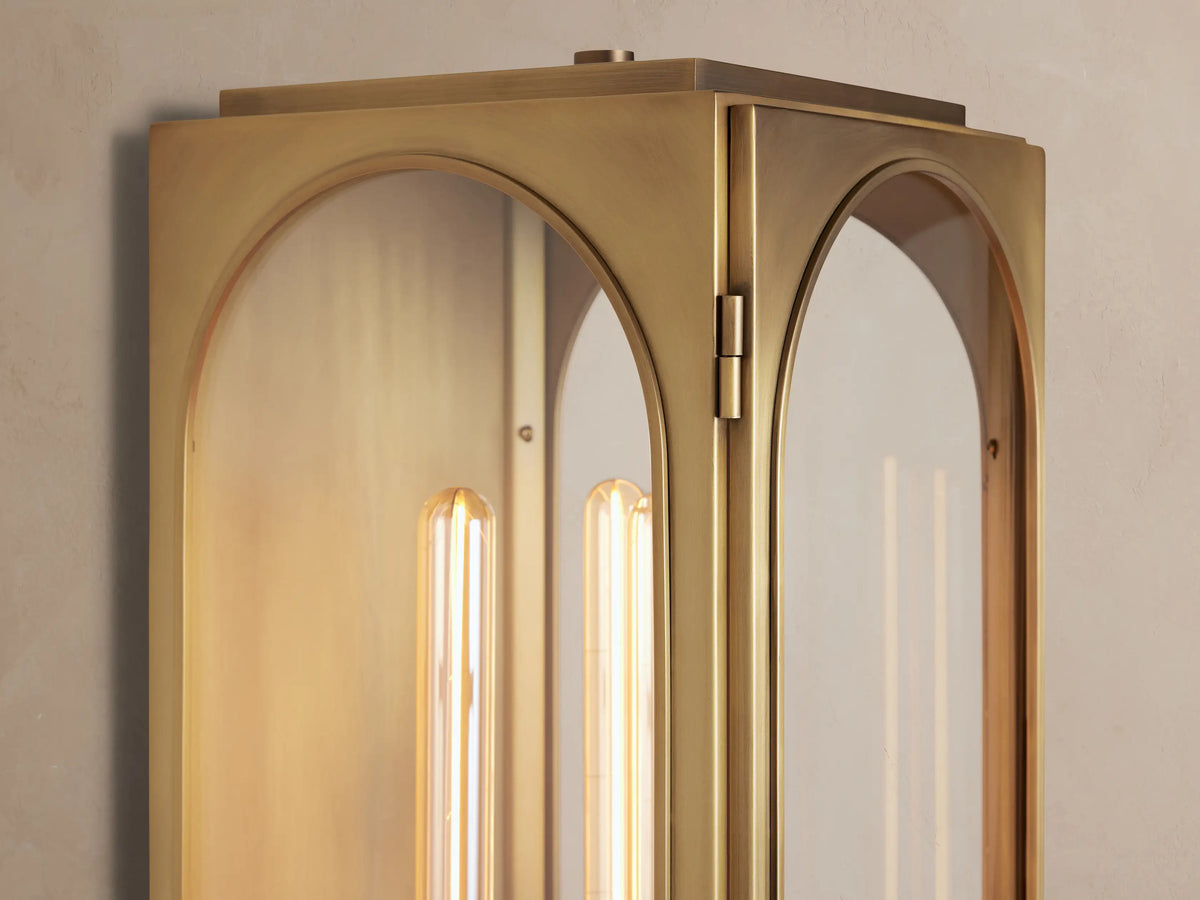 Brass Palladian Outdoor Sconce with a plated finish, clear arched glass, and a vertical linear bulb, mounted on a light wall.