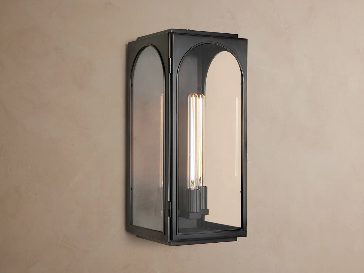 Palladian Outdoor Sconce, dark metal lantern-style wall light with arched glass panels, glowing bulb, on a taupe wall.