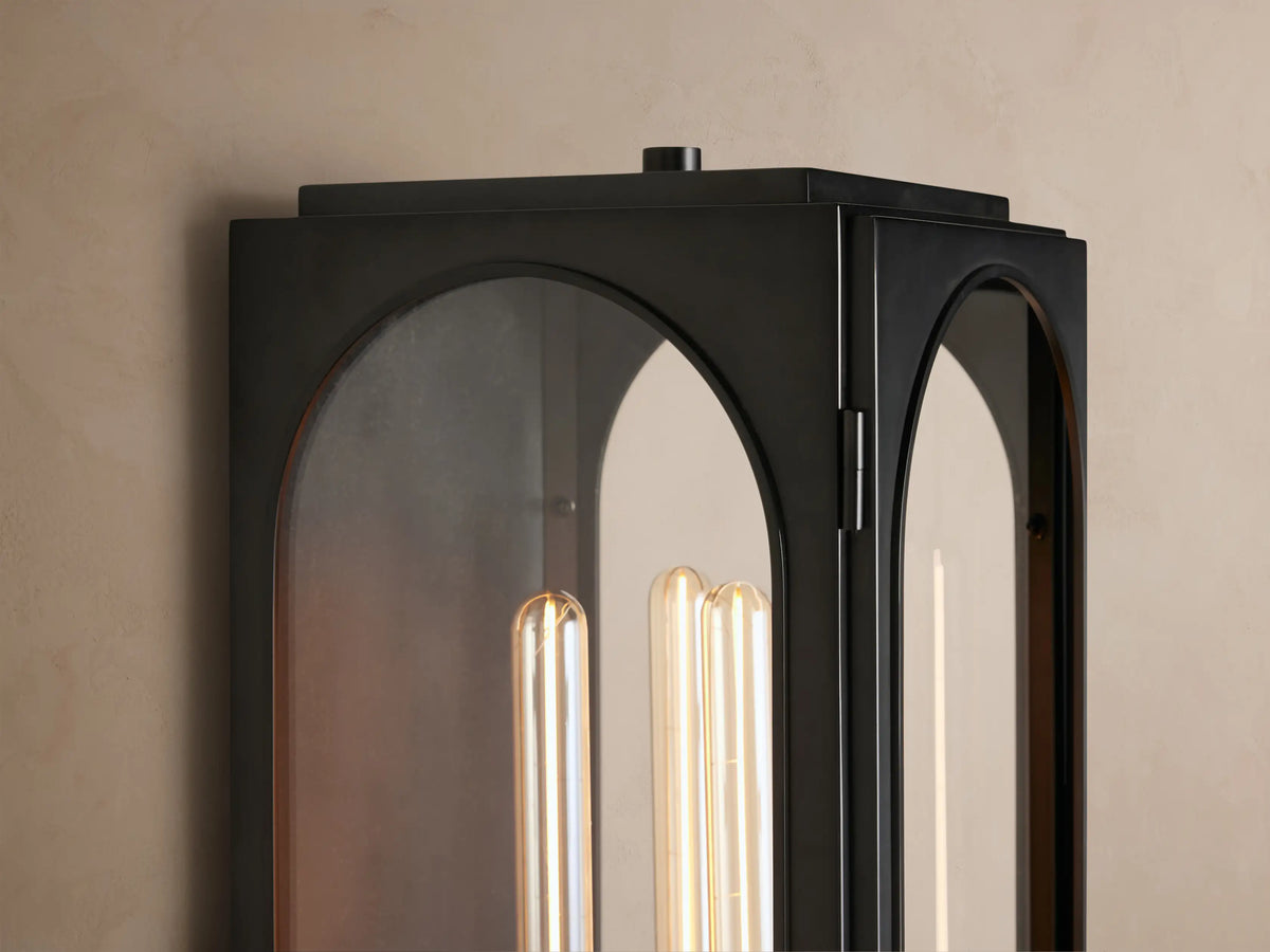 Vertical Palladian Outdoor Sconce, dark metal with two arched clear glass panels, showcasing a glowing filament bulb, on a textured wall.