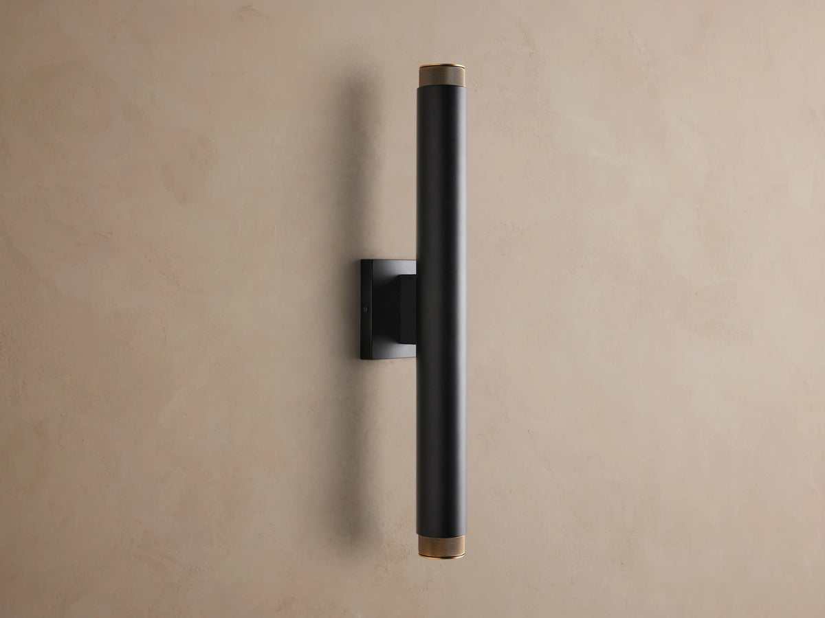 Black Spotlight Outdoor Sconce, vertically mounted, with brass knurled ends, illuminating a textured tan wall.