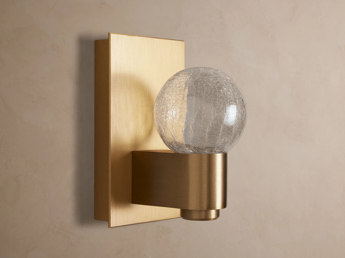 Casen Sconce with satin brass rectangular backplate and spherical crackle glass globe glowing warmly on a wall.
