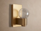 Casen Sconce with satin brass rectangular backplate and spherical crackle glass globe glowing warmly on a wall.