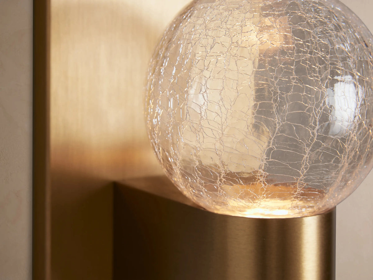 Casen Sconce: Vertical satin brass backplate, arm holding a clear crackle glass globe, mounted on a light wall.