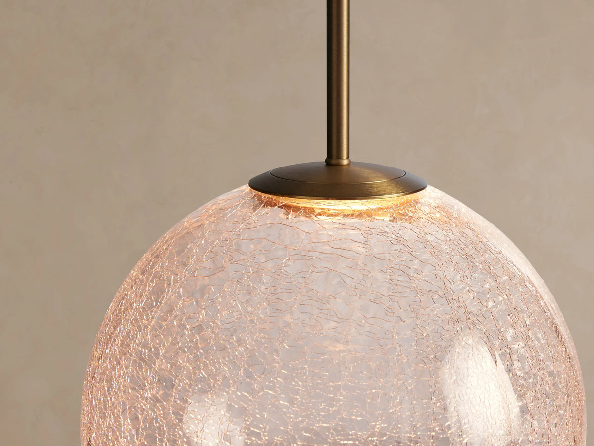 Casen Pendant with a round, crackle glass globe hanging from a satin brass stem and white cord against a beige wall.