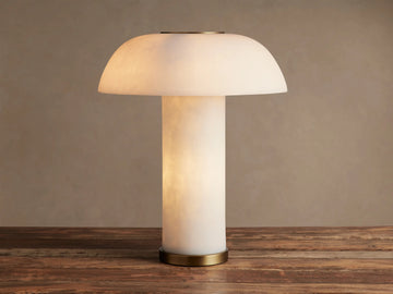 Lit Tramonto Table Lamp, mushroom-shaped, translucent cloudstone resin with brass accents, glowing on a rustic wooden table.