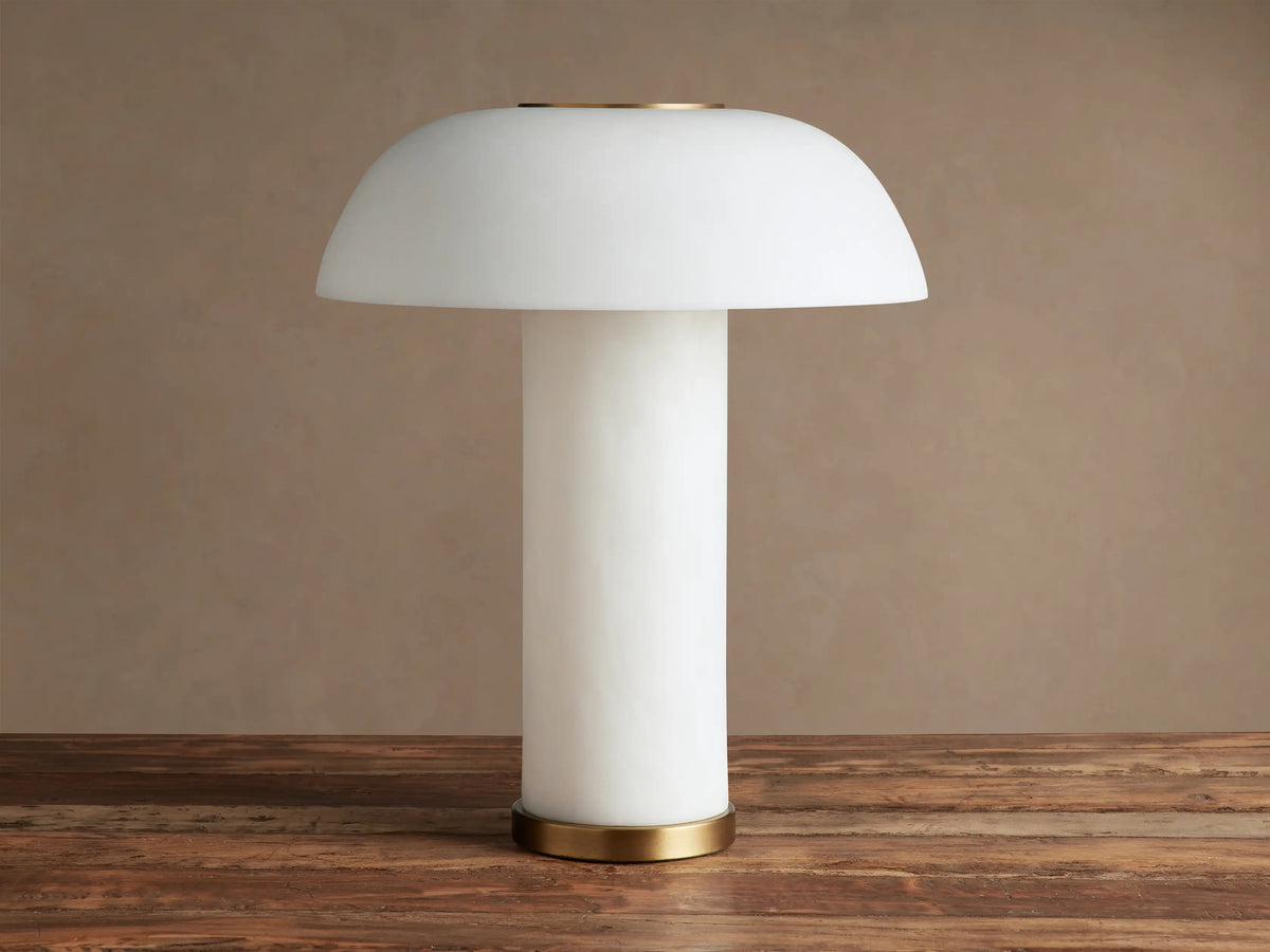 Lit Tramonto Table Lamp, mushroom-shaped, translucent cloudstone resin with brass accents, glowing on a rustic wooden table.