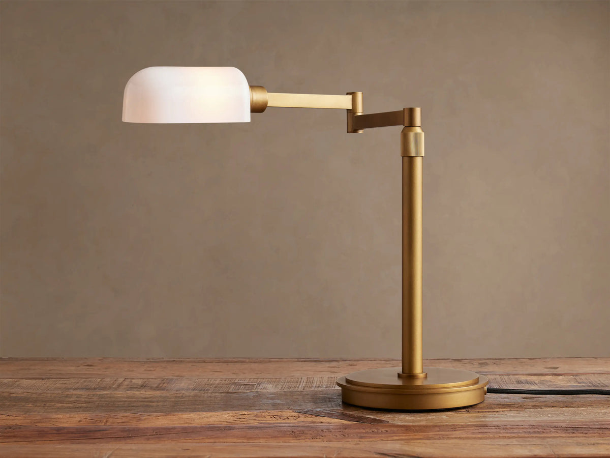 Zayd Swing Arm Table Lamp with brass finish and illuminated white opal glass shade, on a wooden table.