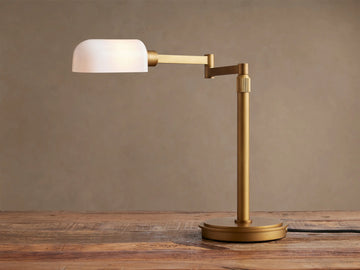 Zayd Swing Arm Table Lamp with brass finish and illuminated white opal glass shade, on a wooden table.
