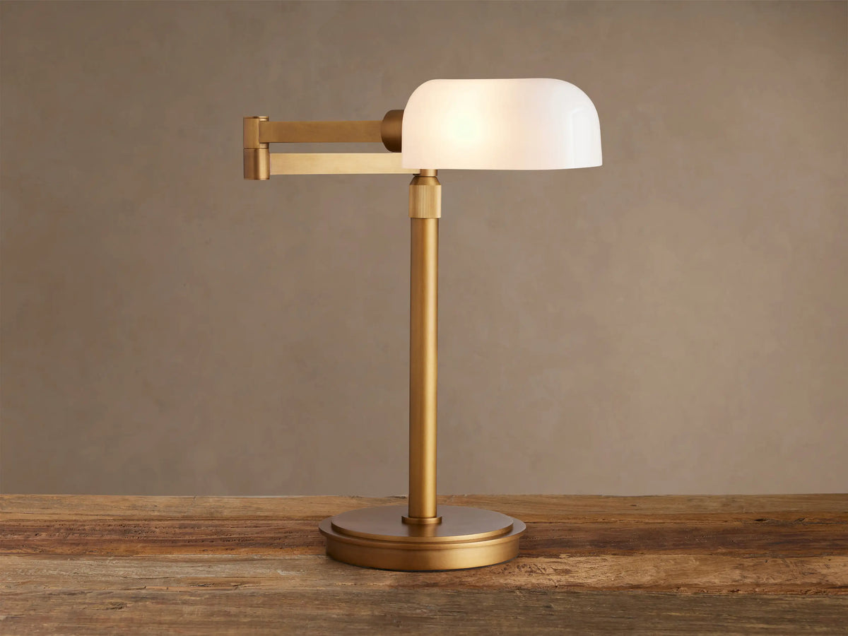 Zayd Swing Arm Table Lamp with brass finish and illuminated white opal glass shade, on a wooden table.