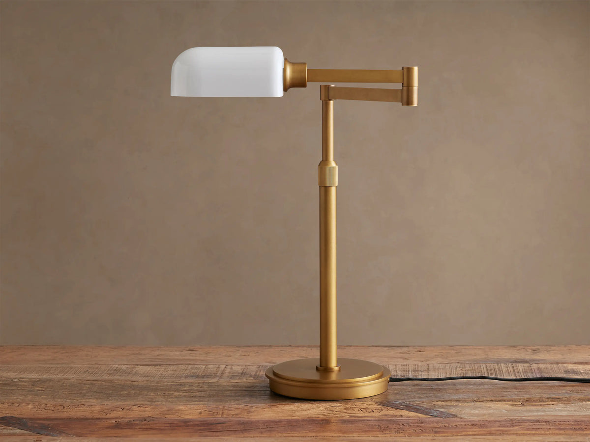 Zayd Swing Arm Table Lamp with brass finish and illuminated white opal glass shade, positioned on a rustic wooden table.