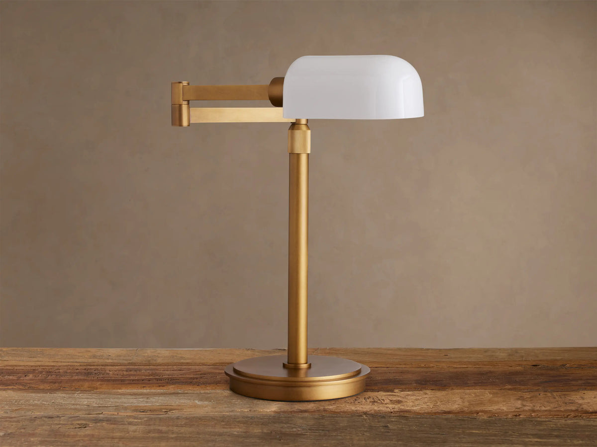 Zayd Swing Arm Table Lamp with brass finish and white opal glass shade, featuring an adjustable arm, on a rustic wooden table.