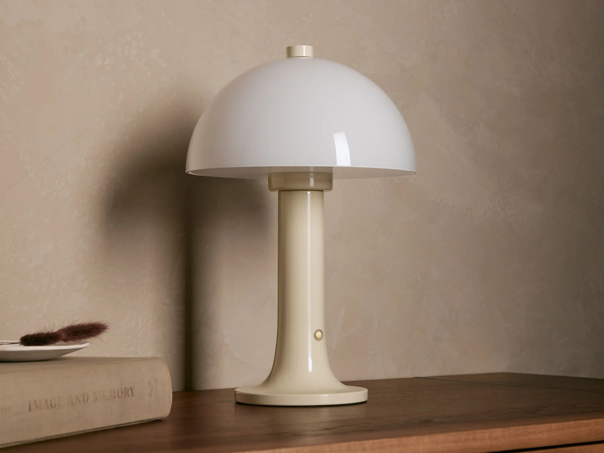 Jocy Portable Table Lamp: Cream metal lamp with a glowing mushroom-shaped opal shade on a wooden table next to a book.