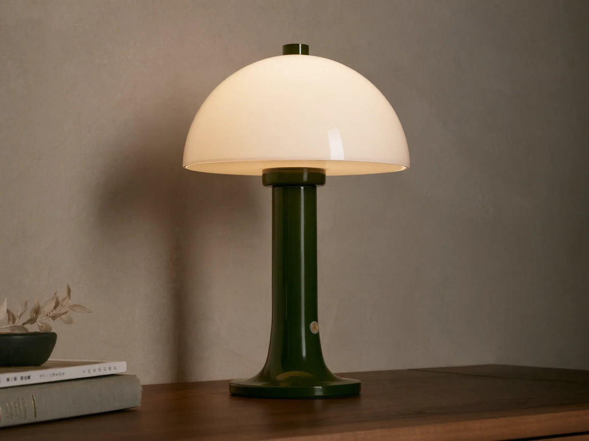 Jocy Portable Table Lamp with a dark green lacquered base and a domed white opal shade, illuminated on a wooden table.