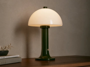 Jocy Portable Table Lamp with a dark green lacquered base and a domed white opal shade, illuminated on a wooden table.