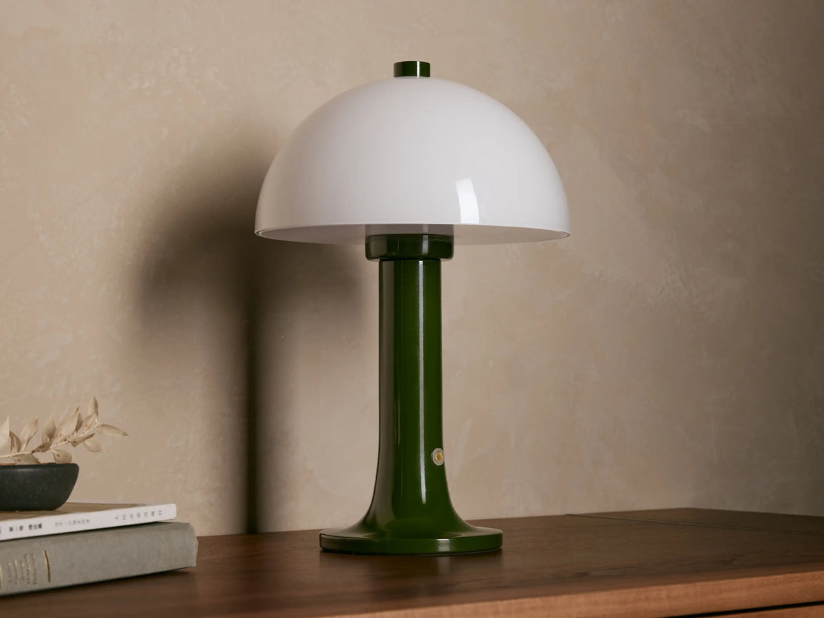 Jocy Portable Table Lamp with a dark green lacquered base and a domed white opal shade, illuminated on a wooden table.