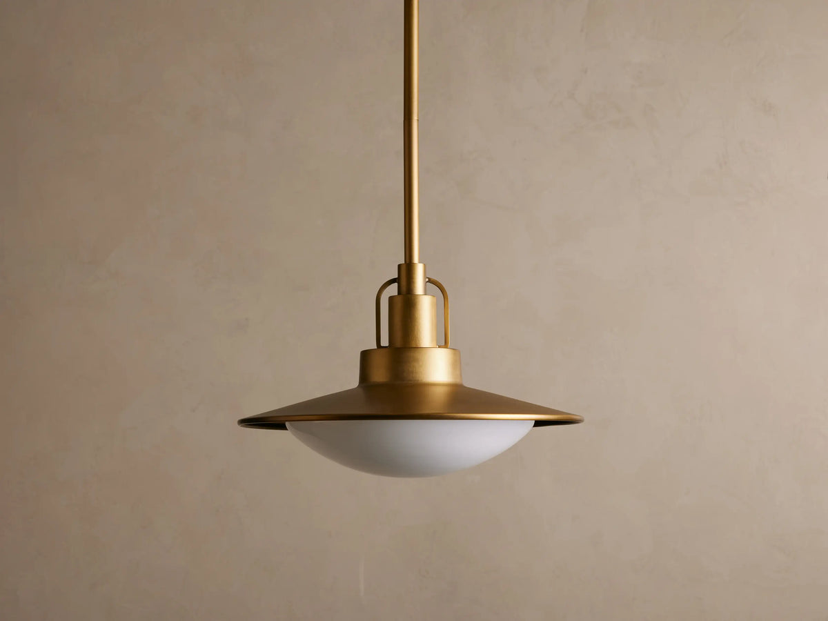 A brass pendant light with a broad, flat circular shade hangs from the ceiling. It emits a warm glow against a neutral backdrop.