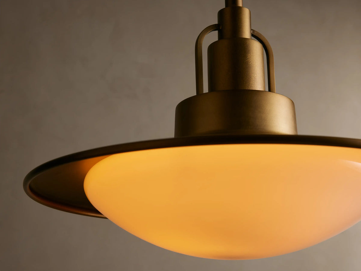 A brass pendant light with a wide, circular, flat shade and frosted white glass diffuser, hanging against a beige wall.