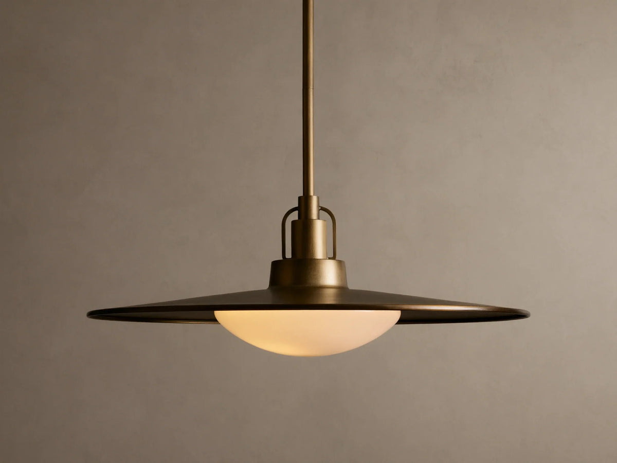 Brass pendant light with a wide, flat shade and a warm glowing bulb. It hangs from a rod against a beige textured wall.