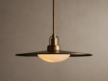 Brass pendant light with a wide, flat shade and a warm glowing bulb. It hangs from a rod against a beige textured wall.