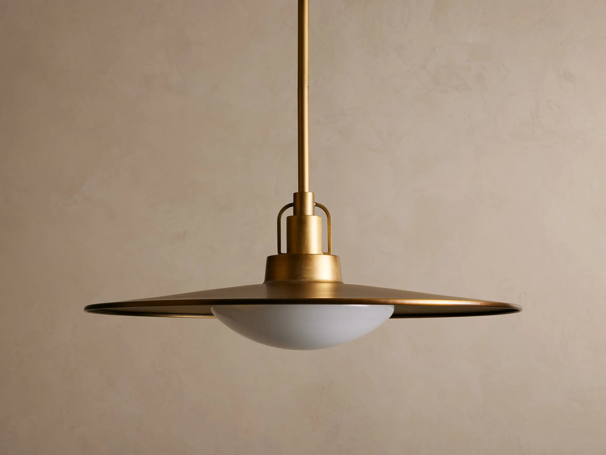 Brass pendant light with a wide, flat shade and a warm glowing bulb. It hangs from a rod against a beige textured wall.