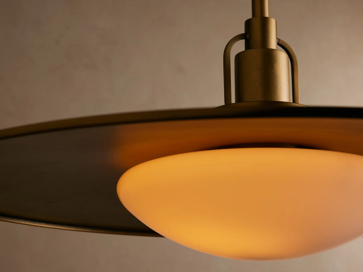 A brass pendant light with a wide, flat shade and frosted white bulb hangs centered against a beige wall.