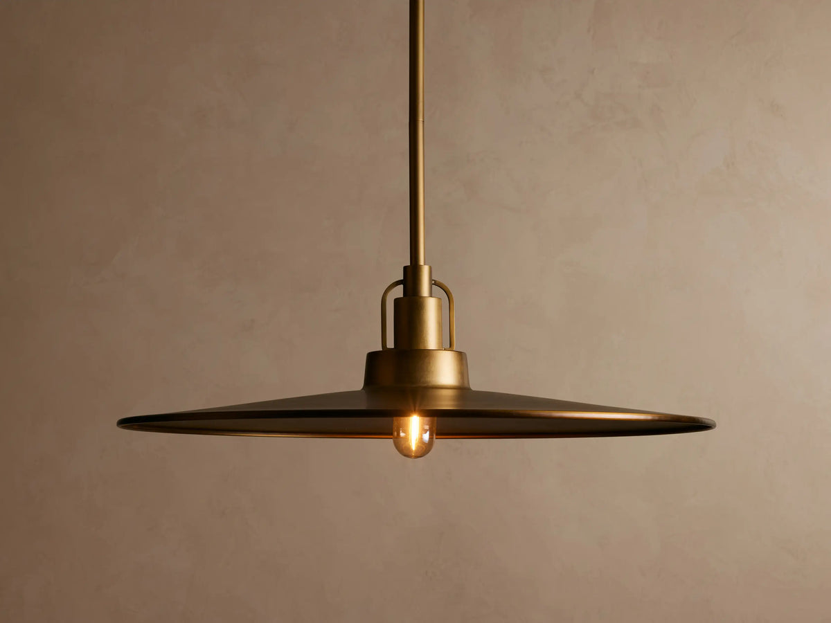 Close-up of a brass pendant light with a flat, round shade, showing a warm metallic finish and a minimalist design against a neutral background.