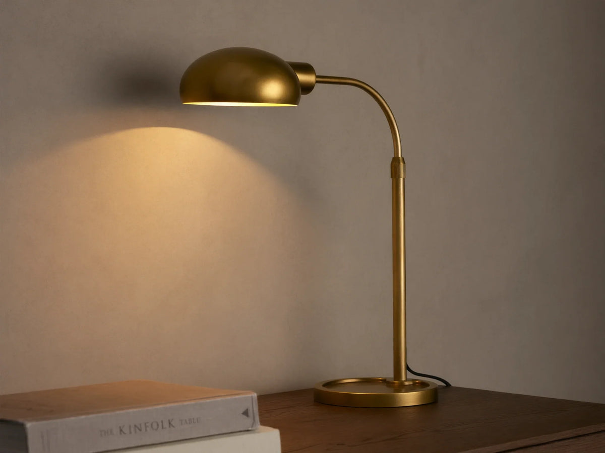 Brass-finished Bergamo Task Table Lamp with a domed shade and curved arm, emitting warm light onto a concrete wall above books on a desk.