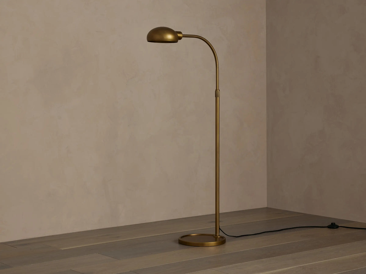 Sleek brass Bergamo Task Floor Lamp with illuminated, pivoting shade, standing on wood floor against a textured wall.