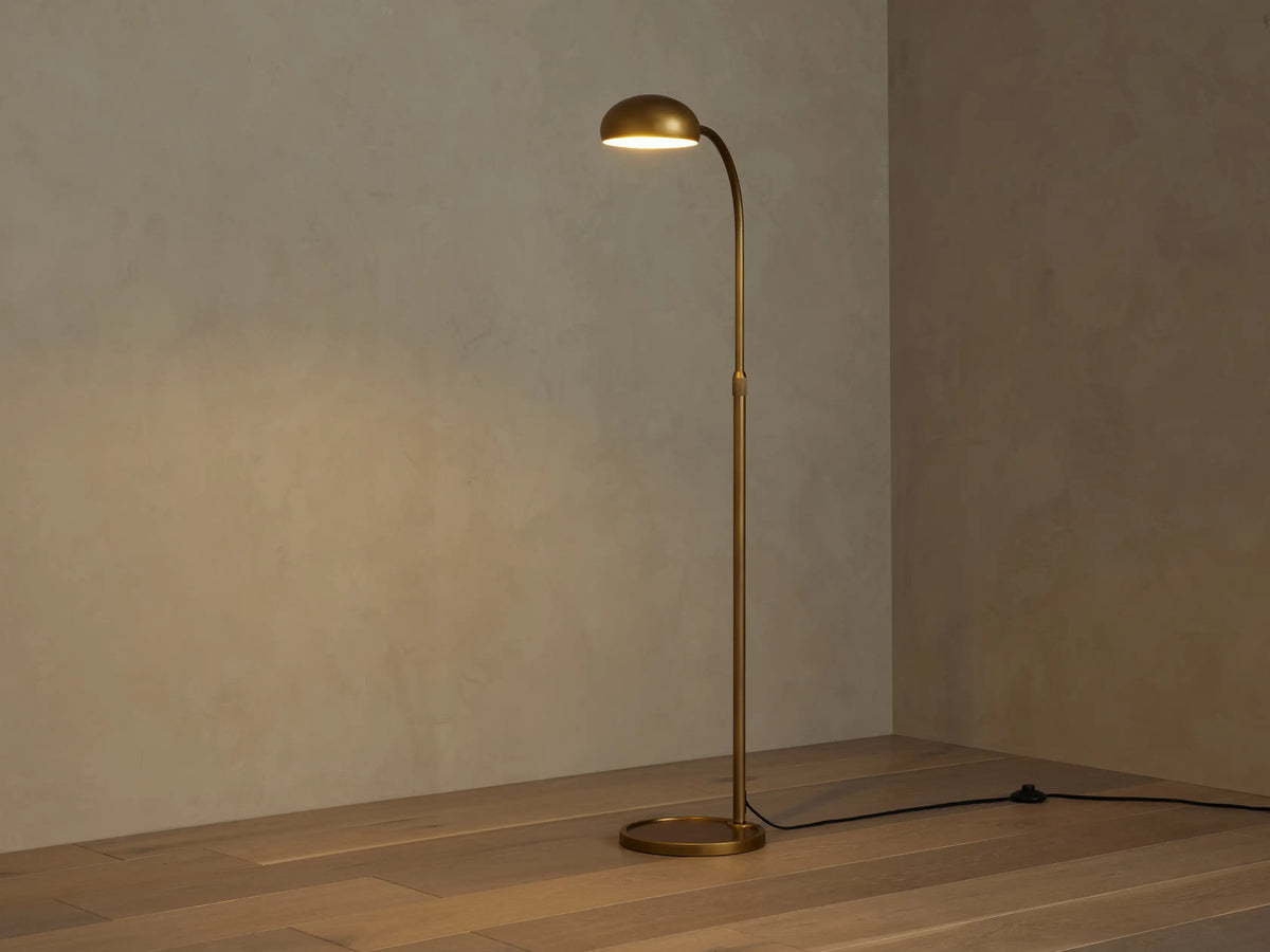 Brass Bergamo Task Floor Lamp with curved arm, round shade, and base on wood floor. Black cord with dimmer switch.