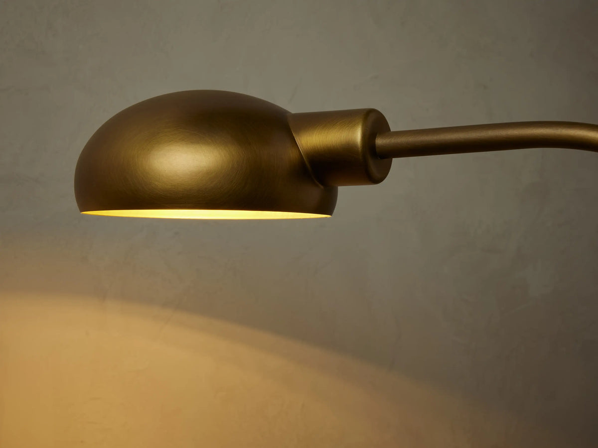 Bergamo Task Floor Lamp, brass finish, curved neck, domed shade with warm light, black cord & dimmer switch on wood floor.