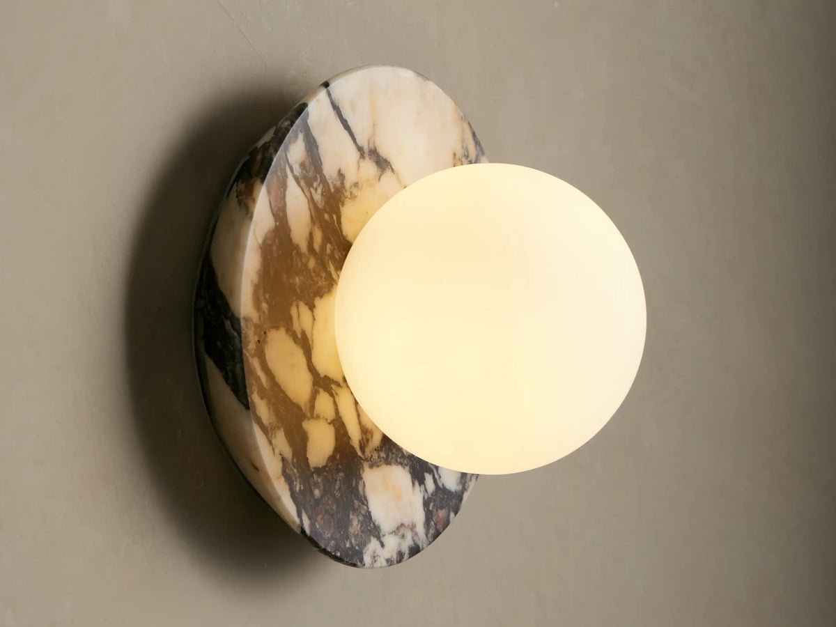 Corinna Sconce: Oval marble backplate with cream, black, and grey veins, featuring a white milk glass globe on a light wall.