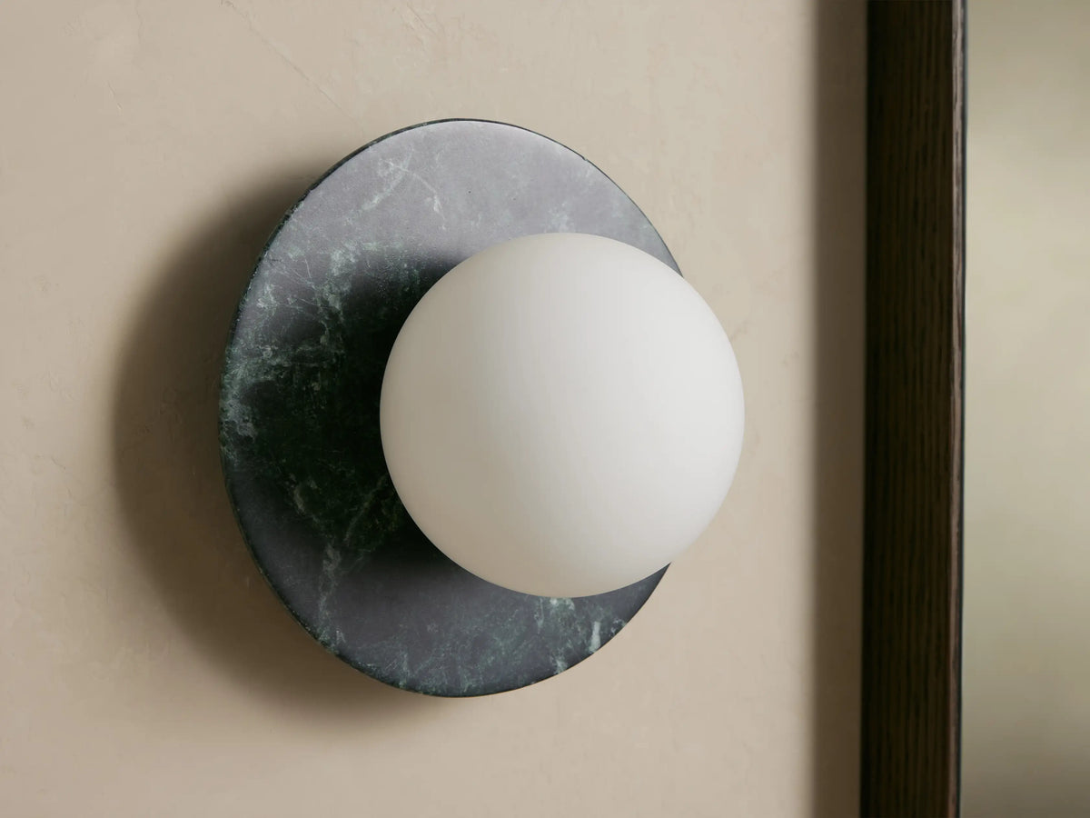 Corinna Sconce: Dark mottled marble circular backplate with an off-center glowing white milk glass globe, mounted on a light wall.