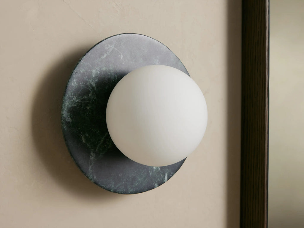 Corinna Sconce: Dark mottled marble circular backplate with an off-center glowing white milk glass globe, mounted on a light wall.