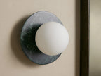 Corinna Sconce: Dark mottled marble circular backplate with an off-center glowing white milk glass globe, mounted on a light wall.
