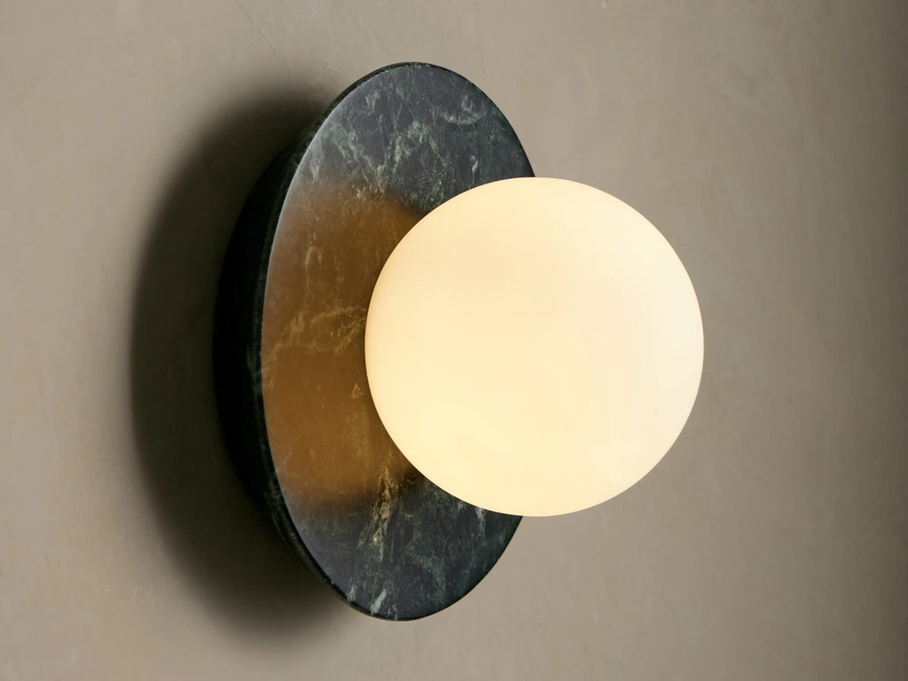Corinna Sconce: Round dark green marble backplate with white veins, supporting a spherical milk glass globe on a beige wall.