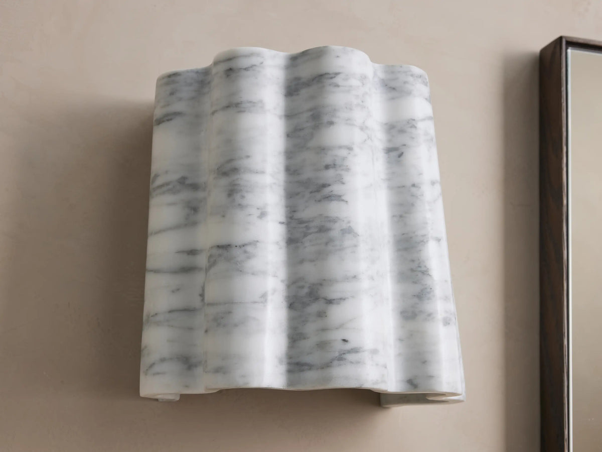 A wall-mounted sconce with a wavy, white marble finish featuring gray veining, installed on a beige wall beside a rectangular mirror.